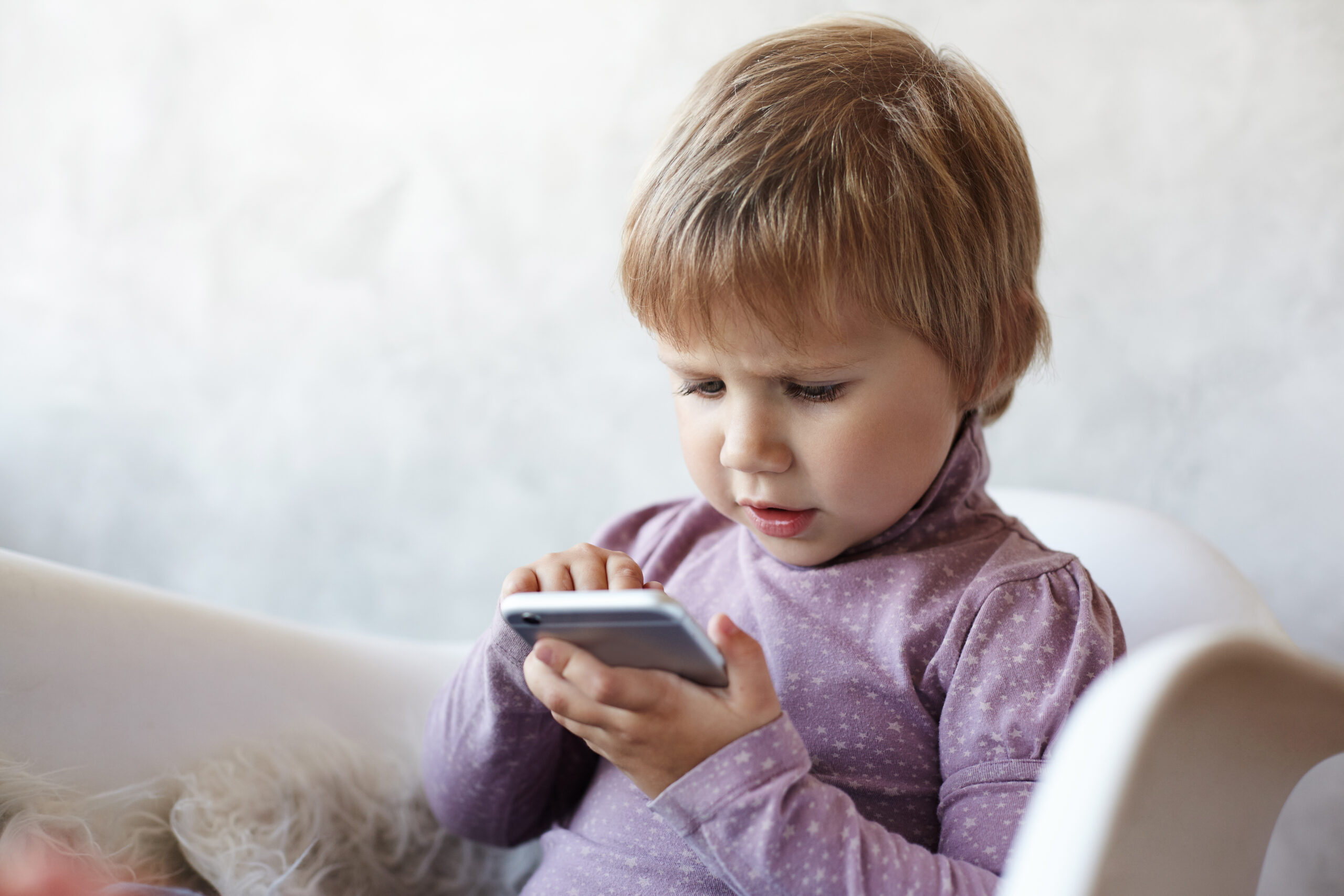 Indoor shot of serious sweet female child with light short hair sitting on armchair using wifi on mobile phone, having focused concentrated look while playing logical game online. Kids and gadgets